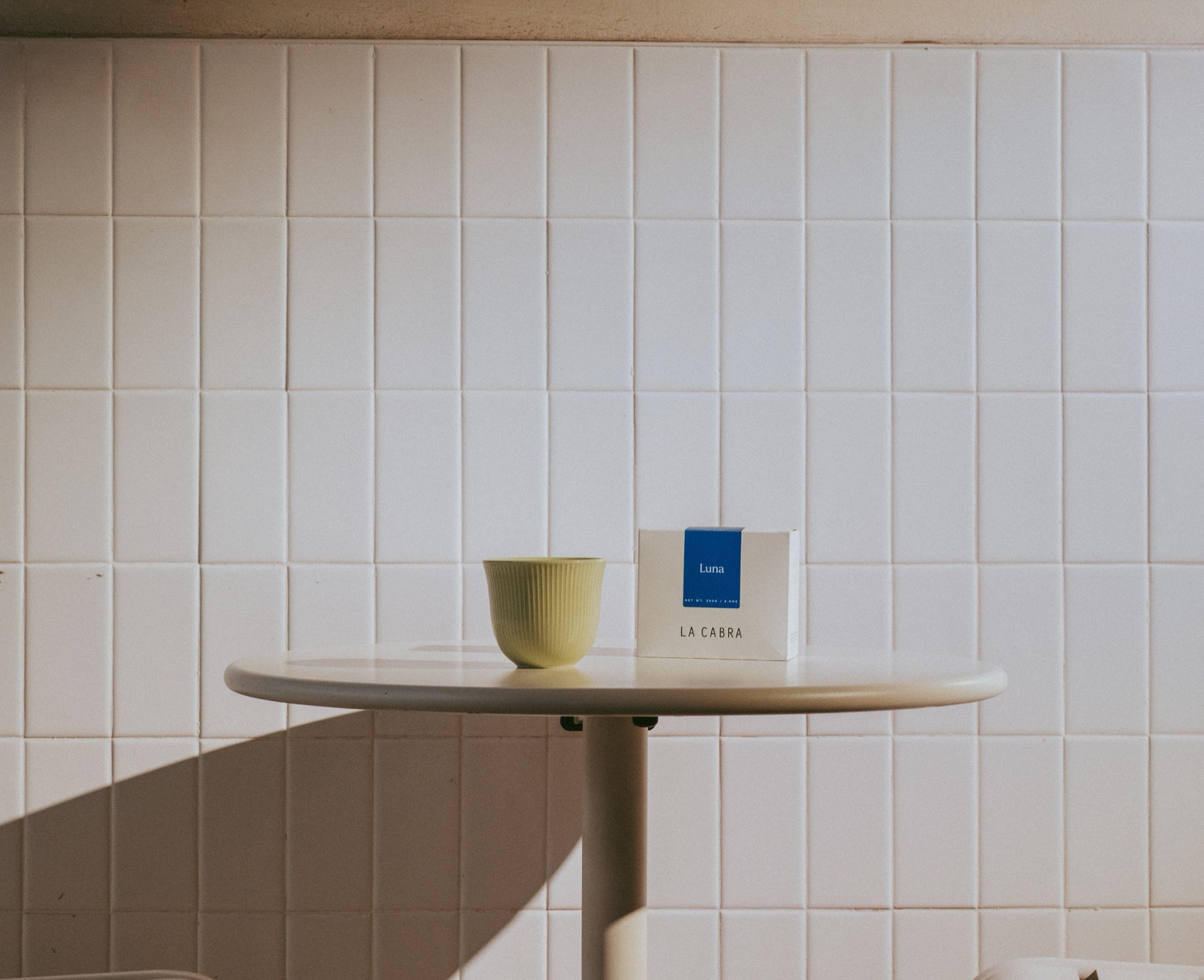 Mesa blanca frente a la pared de losetas con una taza y una caja de café La Cabra